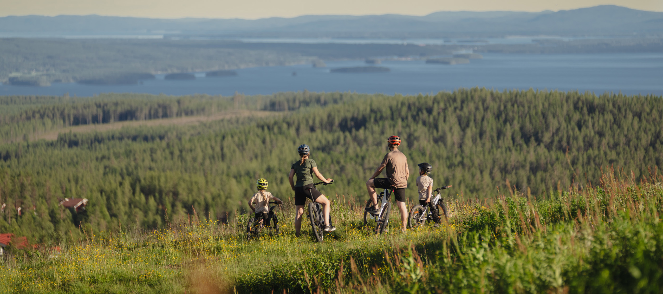 Familj på cyklar blickar ut över skogslandskap och sjöar i kvällsljus.