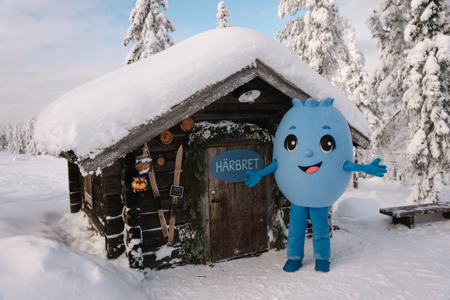 Blå figur står utanför en snötäckt timmerstuga med skylt på dörren i vintermiljö.