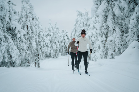 Två personer åker längdskidor på ett preparerat spår genom en snötäckt skog.