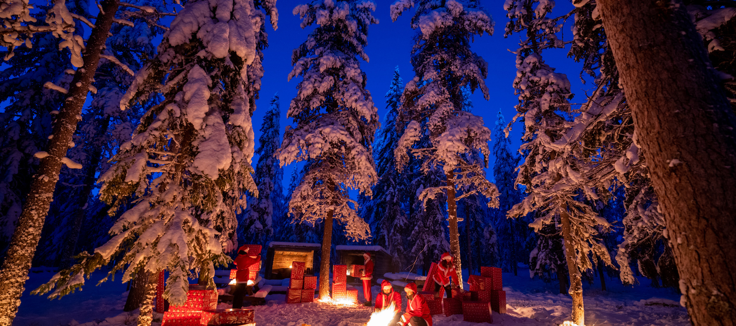 ”Snöklädd vinterkväll i skogen med människor klädda som jultomtar. De sitter vid en brasa och är omgivna av stora inslagna paket och upplysta granar.”
